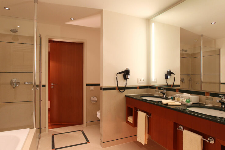 Modernes Hotelbad mit Doppelwaschbecken, Spiegel, Föhn, Dusche und WC. Dezente Beleuchtung und Handtücher vorhanden.