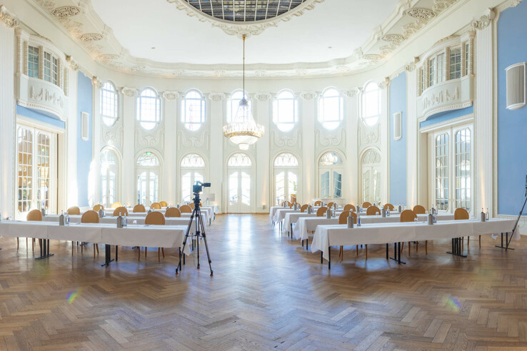 Eleganter, lichtdurchfluteter Konferenzraum mit hohen Decken, blauen Wänden und Kronleuchter, bestuhlt für Veranstaltungen.