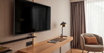 Modernes Hotelzimmer mit Schreibtisch, Lampe, Fernseher und gemütlichem Sessel vor einem Fenster mit Vorhängen.