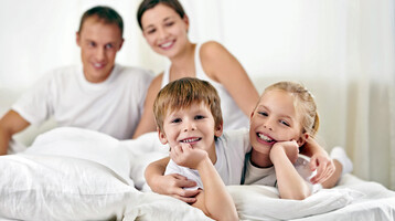Family smiles and relaxes on a hotel bed at the ATLANTIC Hotel Lübeck.