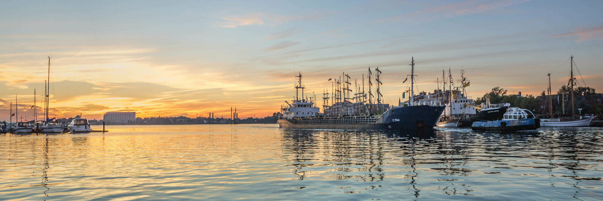 Sunset over a quiet harbor with several sailboats and ships, reflected in the water.