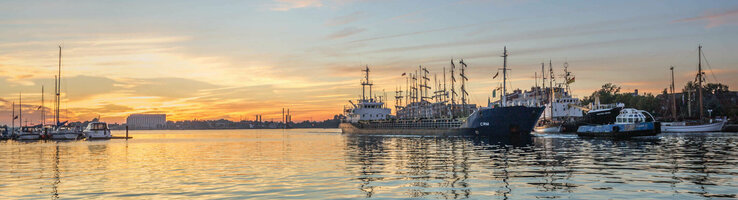 Sunset over a quiet harbor with several sailboats and ships, reflected in the water.
