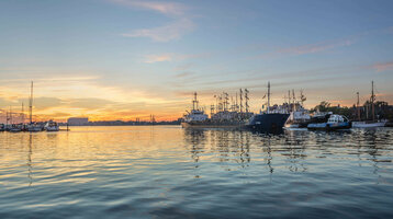 Sunset over a quiet harbor with several sailboats and ships, reflected in the water.
