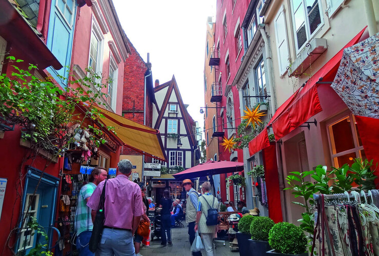 Schmale, belebte Gasse in Bremen mit bunten Häusern, Geschäften und Menschen, die flanieren und einkaufen.