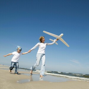 Mutter und Tochter spielen mit einem Flugzeug Mutter und Kind laufen am Strand, sie hält ein Modellflugzeug, blauer Himmel im Hintergrund.