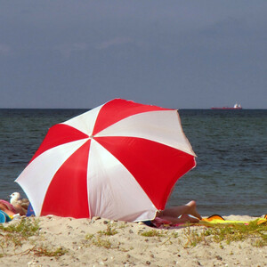 Rot-weißer Sonnenschirm am Sandstrand, dahinter das Meer mit einem Schiff am Horizont.