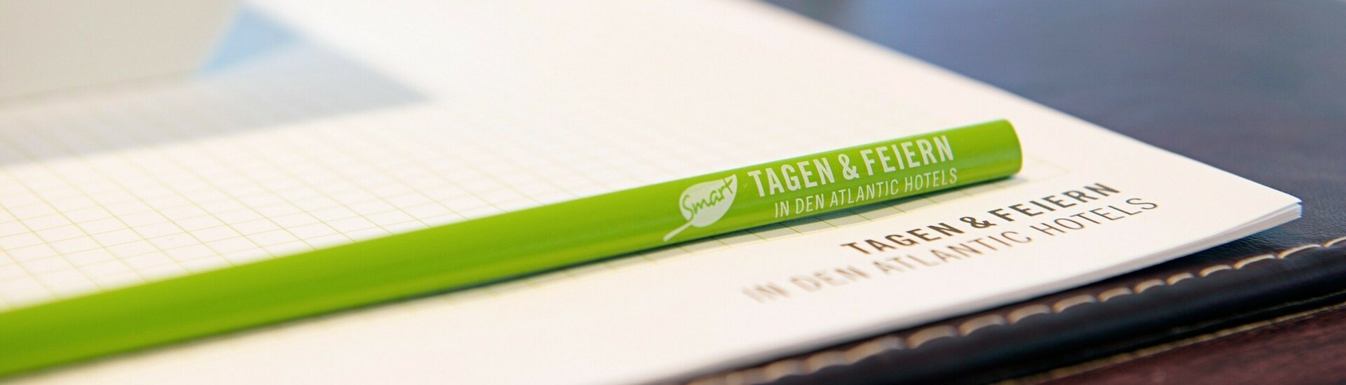 Zettel und Stift im Stil des SMART tagen & feiern Designs auf einem Tisch Grüner Stift und Block mit "Tagen & Feiern" im ATLANTIC Hotel Vegesack, daneben ein Apfel und Wasserglas.