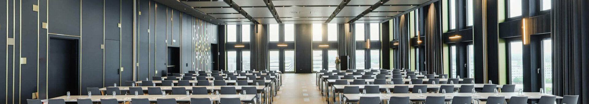 Modern conference room at the ATLANTIC Hotel Heidelberg with rows of tables and chairs, large windows and elegant lighting.