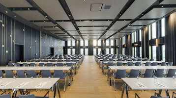 Moderner Konferenzraum im ATLANTIC Hotel Heidelberg mit Reihen von Tischen und Stühlen, großen Fenstern und eleganter Beleuchtung.