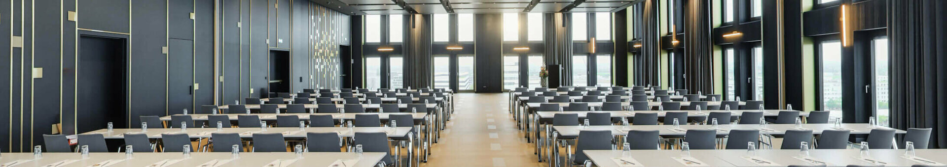 Moderner Konferenzraum im ATLANTIC Hotel Heidelberg mit Reihen von Tischen und Stühlen, großen Fenstern und eleganter Beleuchtung.