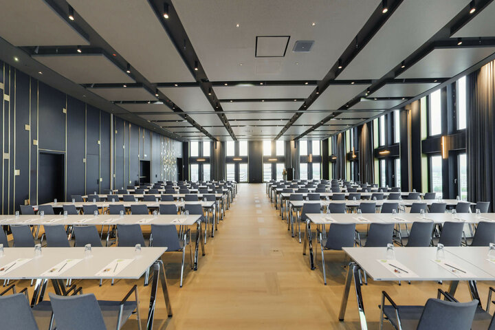 Moderner Konferenzraum im ATLANTIC Hotel Heidelberg mit Reihen von Tischen und Stühlen, großen Fenstern und eleganter Beleuchtung.