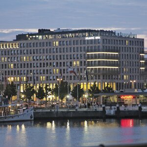 Modern hotel on the waterfront at dusk, illuminated, with a ship in the foreground and restaurants along the promenade.