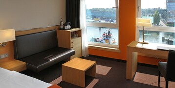 Hotelzimmer mit Hafenblick: modernes Interieur, Sofa, Schreibtisch, große Fenster mit Blick auf Wasser und Schiffe.