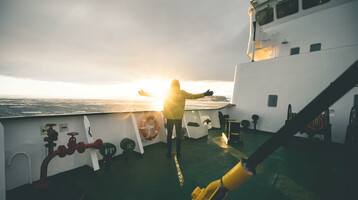 Film-Projekt Antarktis | Sponsoring durch ATLANTIC Hotel Sail City Bremerhaven Person auf Schiffdeck genießt Sonnenuntergang, mit ausgestreckten Armen, Meer im Hintergrund.