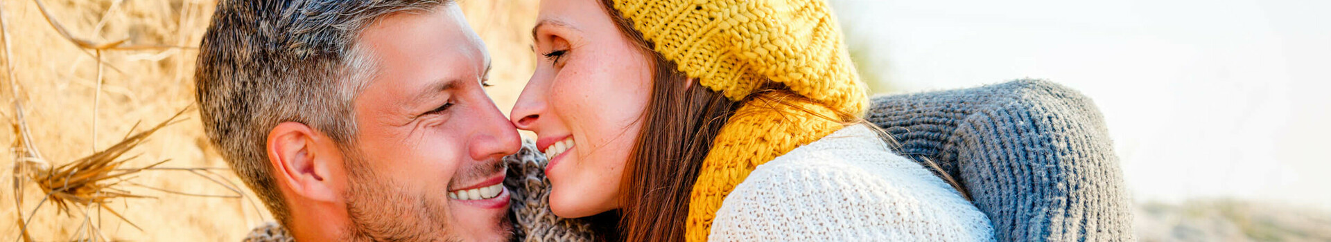 Paar in warmer Kleidung kuschelt am Strand, lächelt sich an, entspannte Atmosphäre.