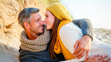 Paar in warmer Kleidung kuschelt am Strand, lächelt sich an, entspannte Atmosphäre.
