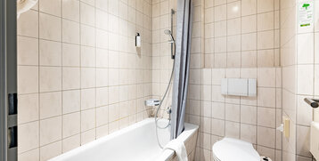Helles Badezimmer mit Badewanne, Duschvorhang und Toilette im ATLANTIC Hotel Vegesack, Bremen.