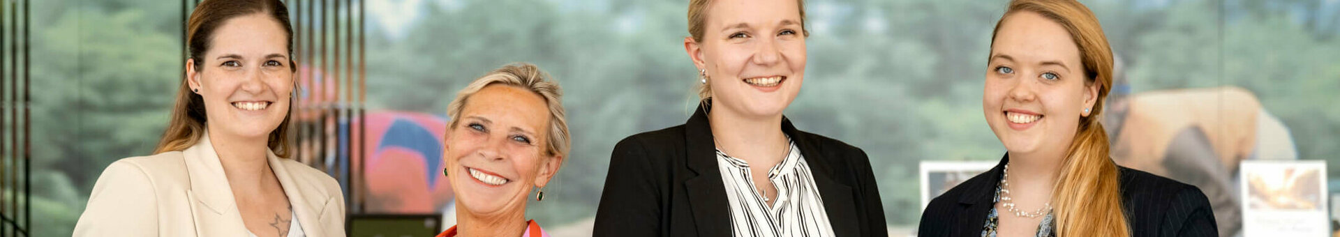 Vier lächelnde Frauen in Businesskleidung vor einem Hintergrund mit Naturmotiv im ATLANTIC Hotel Galopprennbahn.