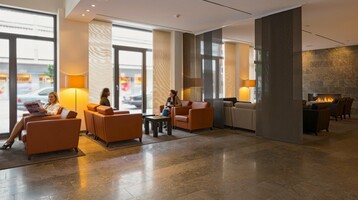 Bright hotel lobby with leather armchairs, fireplace, large windows and guests relaxing and chatting.