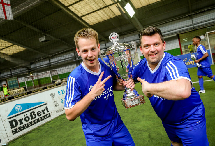 Zwei Männer im ATLANTIC Hotel-Trikot halten jubelnd einen großen Pokal in einer Sporthalle.
