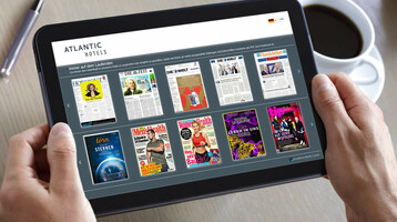 Tablet shows digital magazine selection in a hotel, held by hands, next to a cup of coffee and a pen.