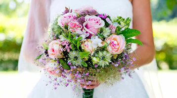 Braut hält einen üppigen Blumenstrauß mit rosa Rosen und grünen Akzenten vor einem verschwommenen, grünen Hintergrund.