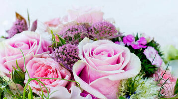 Braut hält einen üppigen Blumenstrauß mit rosa Rosen und grünen Akzenten vor einem verschwommenen, grünen Hintergrund.