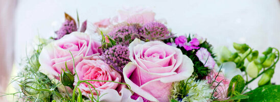 Braut hält einen üppigen Blumenstrauß mit rosa Rosen und grünen Akzenten vor einem verschwommenen, grünen Hintergrund.