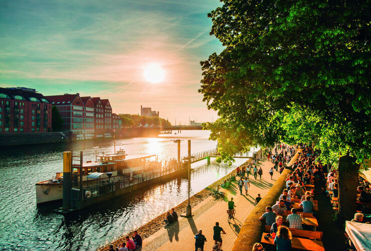 Uferpromenade in Bremen bei Sonnenuntergang, mit Menschen, Bäumen und einem Boot am Fluss.