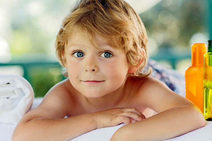 Kind mit blonden Haaren liegt lächelnd auf einem Handtuch neben Sonnencremeflaschen.