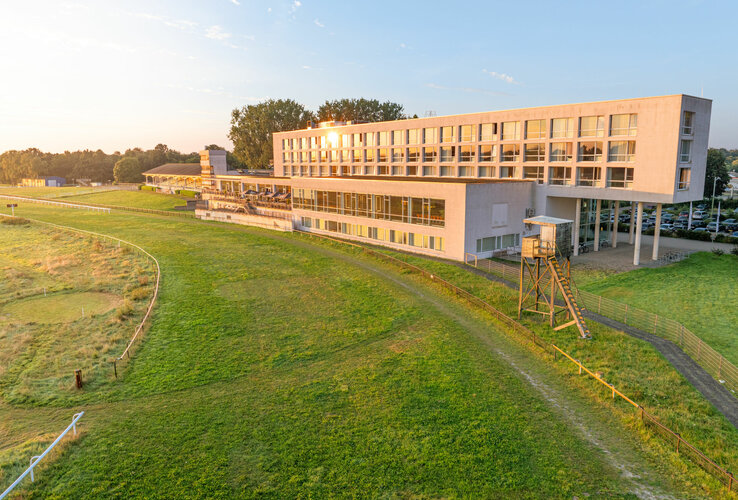 Modernes Hotelgebäude am Rande einer grünen Rennbahn, umgeben von Bäumen und beleuchtet von der Abendsonne.