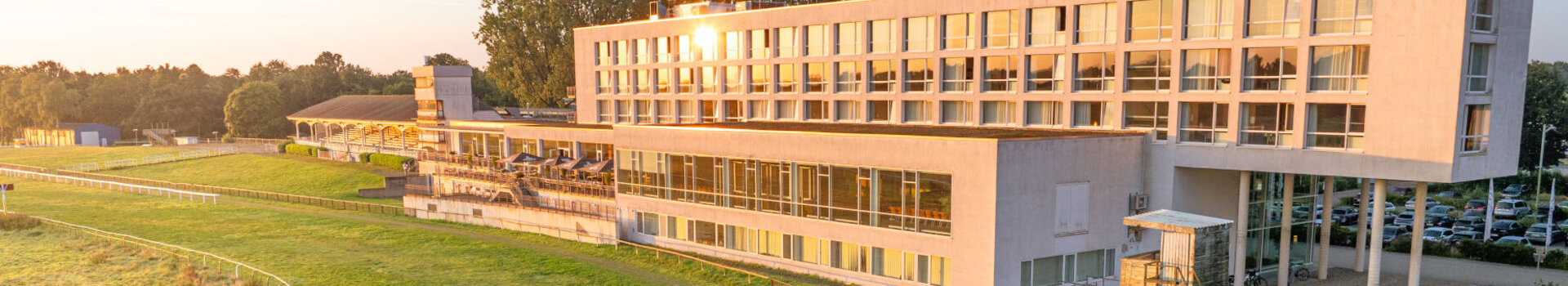 Modernes Hotelgebäude am Rande einer grünen Rennbahn, umgeben von Bäumen und beleuchtet von der Abendsonne.