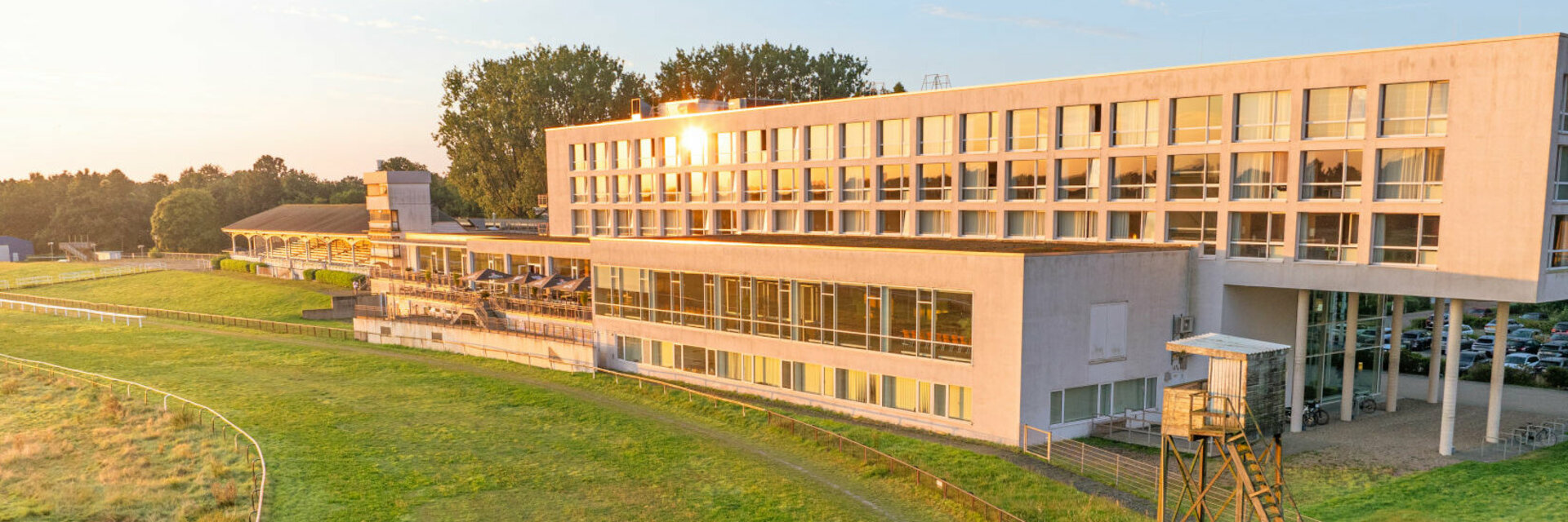Modern hotel building on the edge of a green racecourse, surrounded by trees and illuminated by the evening sun.