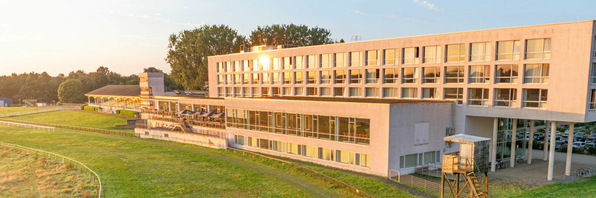 Modernes Hotelgebäude am Rande einer grünen Rennbahn, umgeben von Bäumen und beleuchtet von der Abendsonne.