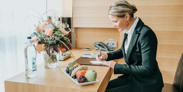 Frau im Anzug schreibt an einem Schreibtisch im Hotelzimmer, mit Blumen, Wasserflasche und Obstschale.