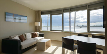 Bright hotel room with sofa, dining table and large window with a view of the water and the city in Bremerhaven.