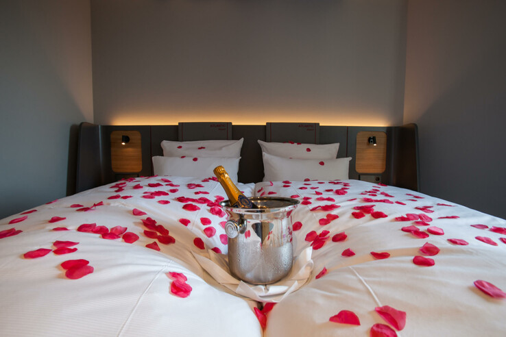 Hotel room with double bed, red rose petals and champagne cooler on white bed linen.