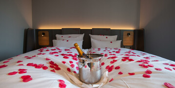 Hotelzimmer mit Doppelbett, roten Rosenblättern und Sektkühler auf weißer Bettwäsche.