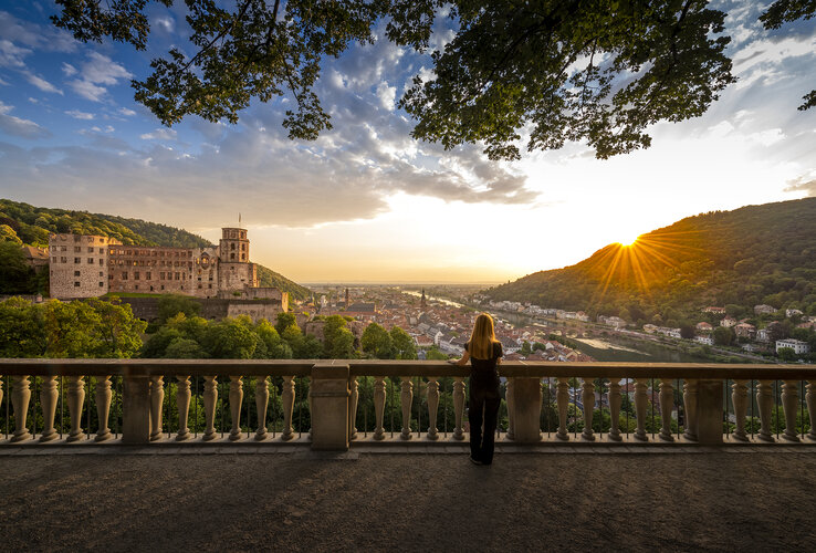 Frau blickt von Terrasse auf Heidelberger Schloss und Stadt im Sonnenuntergang.