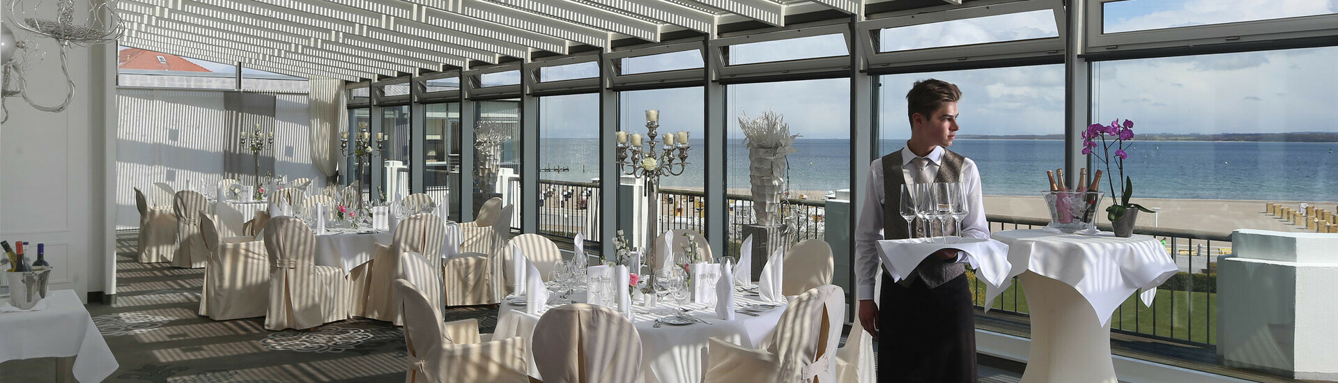 Elegant hotel restaurant with a sea view, laid tables and a waiter carrying glasses.