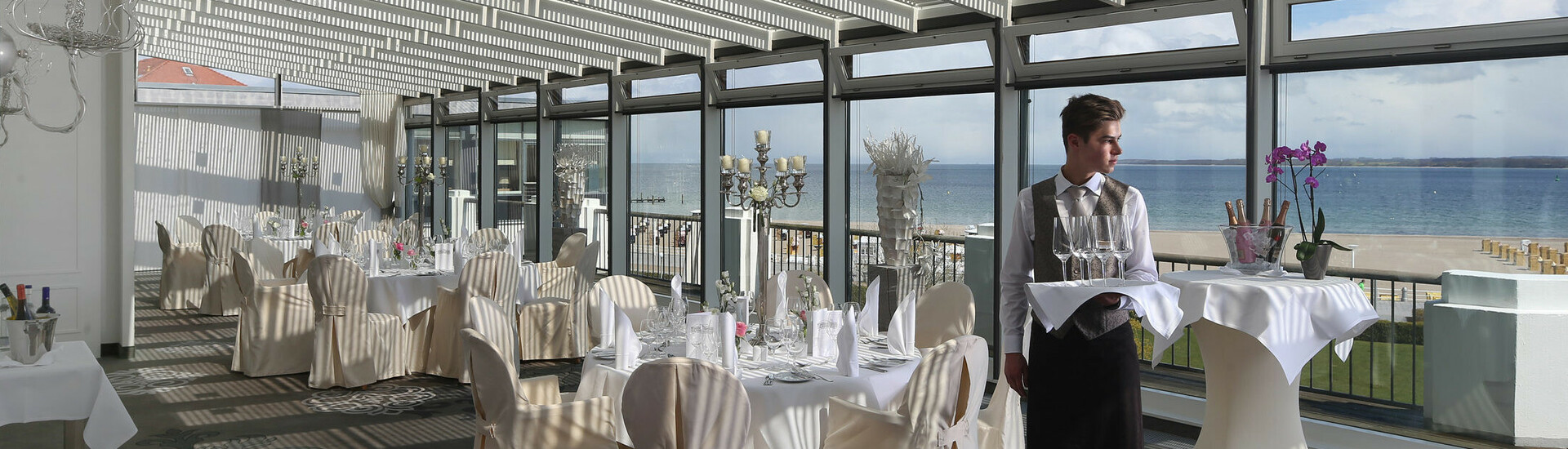 Elegantes Hotelrestaurant mit Meerblick, gedeckten Tischen und einem Kellner, der Gläser trägt.