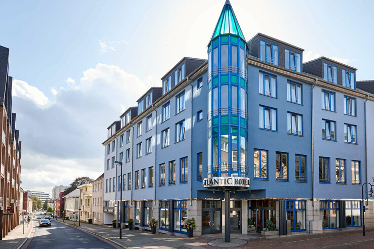 Außenansicht des ATLANTIC Hotel Vegesack in Bremen, modernes Gebäude mit blauer Glasfassade und Straßenszene.