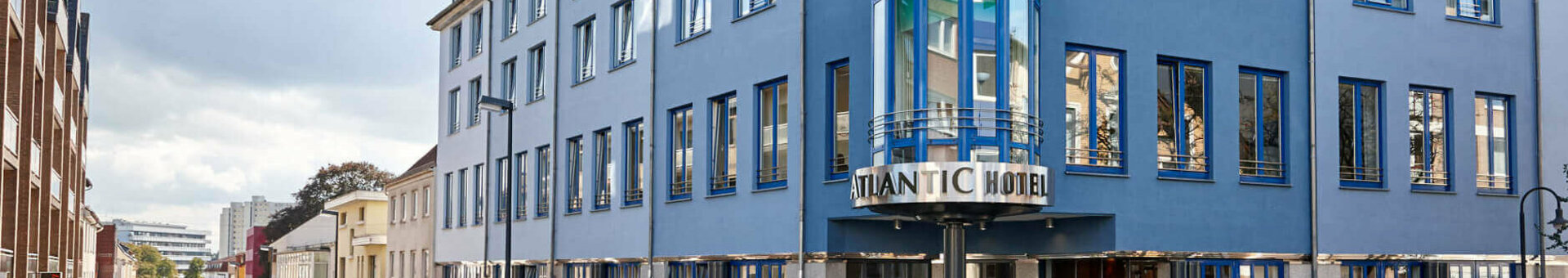 Außenansicht des ATLANTIC Hotel Vegesack in Bremen, modernes Gebäude mit blauer Glasfassade und Straßenszene.