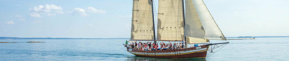 Sailing ship with white sails on a calm sea under a blue sky, full of passengers.