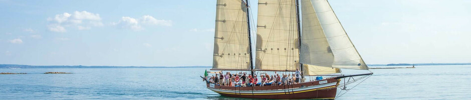 Segelschiff mit weißen Segeln auf ruhiger See unter blauem Himmel, voller Passagiere.