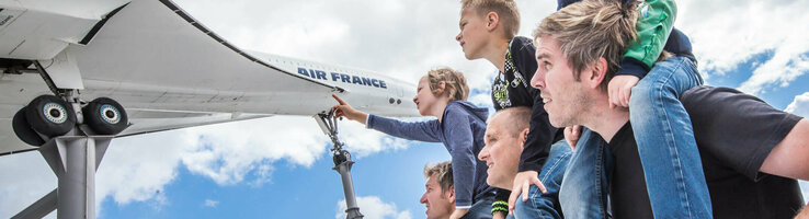 Family looking at airplane model outdoors, children on shoulders, blue sky in the background.