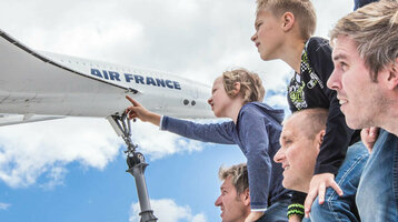 Family looking at airplane model outdoors, children on shoulders, blue sky in the background.