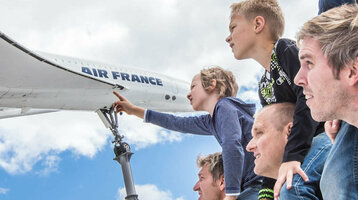 Familie betrachtet Flugzeugmodell im Freien, Kinder auf Schultern, blauer Himmel im Hintergrund.