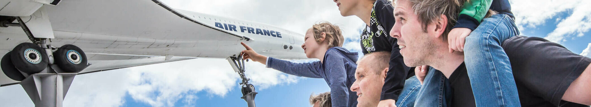 Familie betrachtet Flugzeugmodell im Freien, Kinder auf Schultern, blauer Himmel im Hintergrund.
