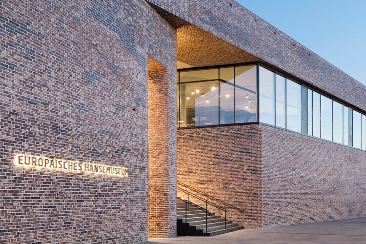Backsteingebäude des Europäischen Hansemuseums mit beleuchtetem Eingang und großen Fenstern bei Abenddämmerung.